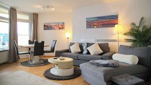 Ein modernes Wohnzimmer mit einem grauen Sofa, einem runden Couchtisch, einem Esstisch mit Stühlen und einem großen Fenster mit Vorhängen.