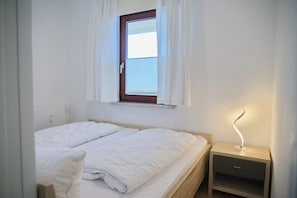 A bedroom with a bed, a nightstand, a lamp, and a window with curtains. - Grömitz-center I App. 648 (Grömitz)