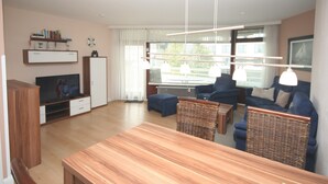 En moderne stue med et træbord, blå sofaer, et fjernsyn og et stort vindue med gardiner.
