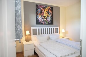 A hotel room with a bed, two bedside tables, a nightstand, a wall-mounted painting, and a door with a patterned curtain.