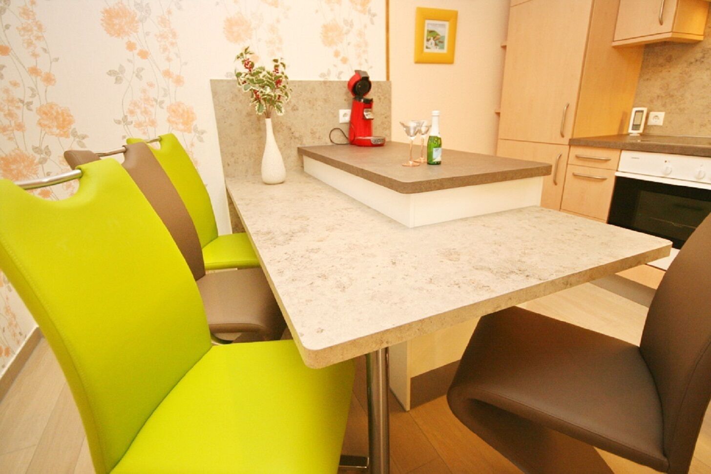 A compact kitchen with a countertop, a coffee machine, a vase with flowers, and a bottle of wine.