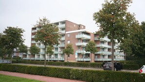 Un immeuble résidentiel à plusieurs étages, doté de balcons, d’une allée pavée et de voitures garées.