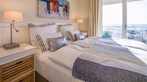 Quarto de hotel com uma cama grande, duas mesas de cabeceira com luminárias, um quadro na parede e vista para o mar através de uma grande janela.