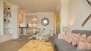 Uma sala de estar moderna com um sofá cinza, almofadas coloridas listradas, uma mesa de centro de madeira e um espaço de jantar com cadeiras de vime.