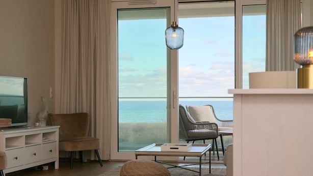 A modern living room with a television, a sofa, a chair, a coffee table, and a view of the ocean through large glass doors.