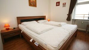 A neatly made bed with a wooden headboard, a bedside table with a lamp, and a window with curtains. - Prinzenhof App. 26 (Grömitz)