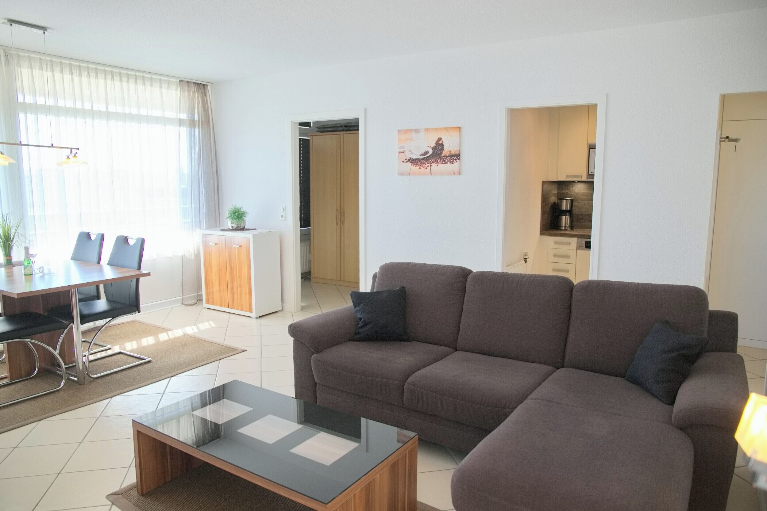 A modern living room with a brown sofa, a glass coffee table, and a dining area with a table and chairs. There is a kitchen area visible through an open door.