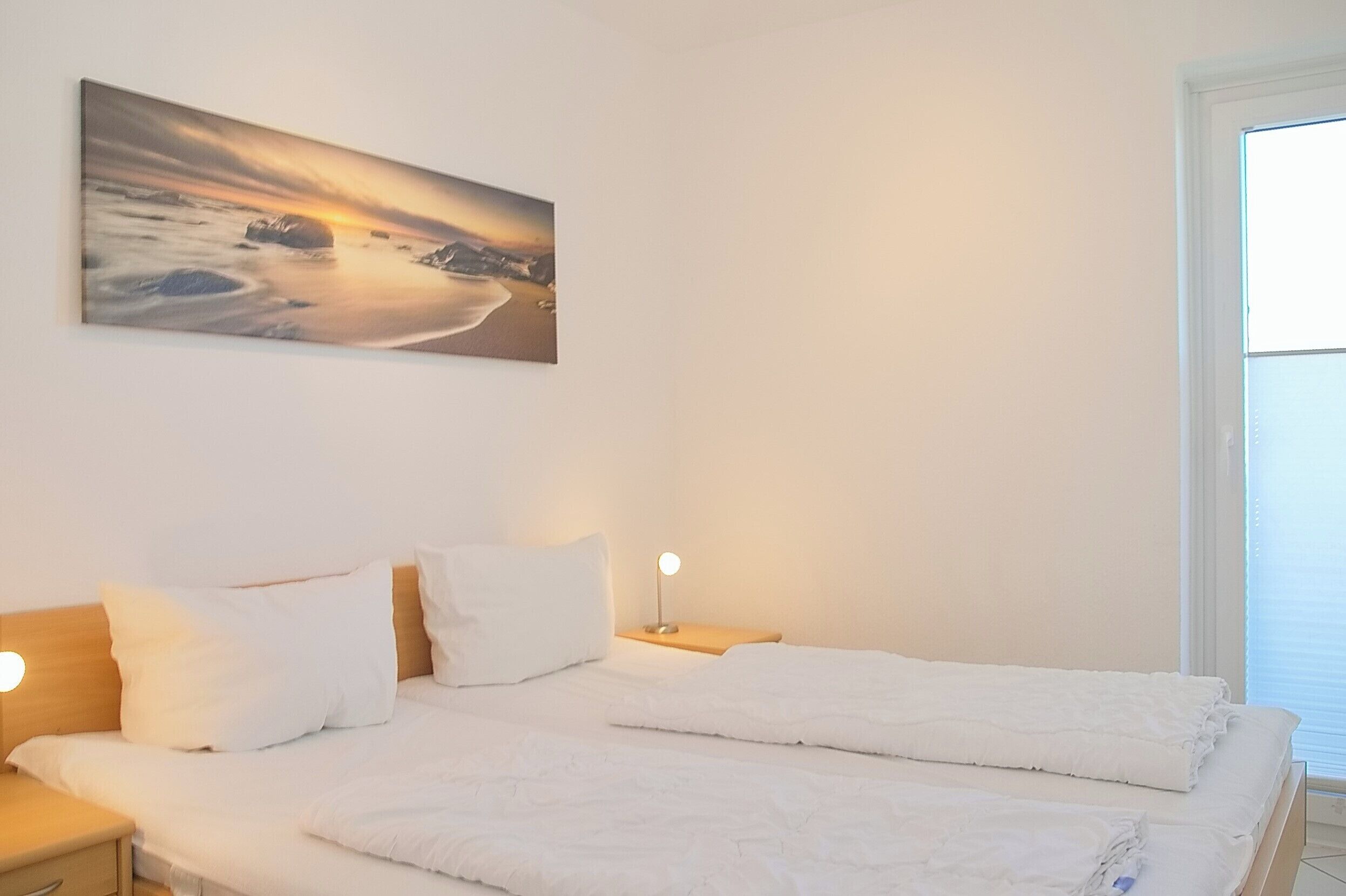 Ein ordentlich bezogenes Bett mit weißen Leinen, ein Nachttisch mit Lampe und ein Gemälde mit Sonnenuntergang über einem Strand an der Wand.