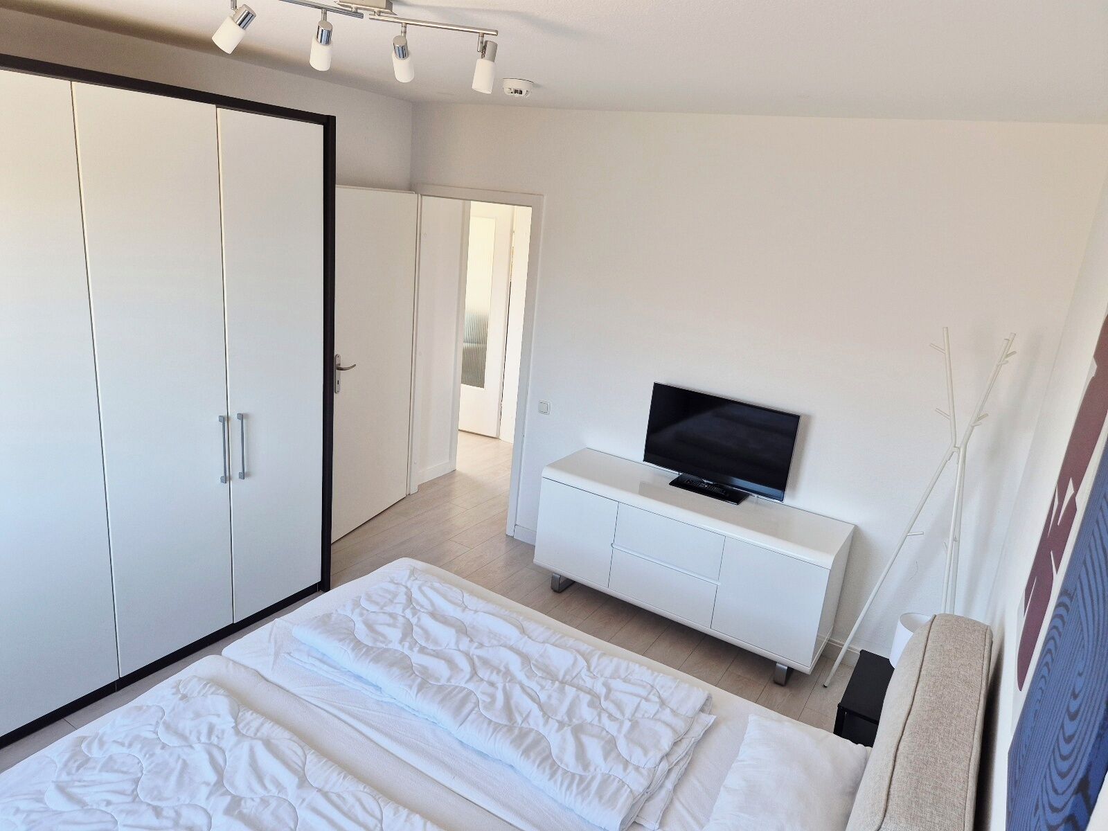A bedroom with a bed, a TV on a white cabinet, a wardrobe, and a door leading to another room.