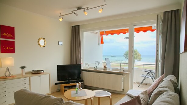 Een woonkamer met een bank, televisie en een tafel met een fles. Er is een raam met uitzicht op zee.