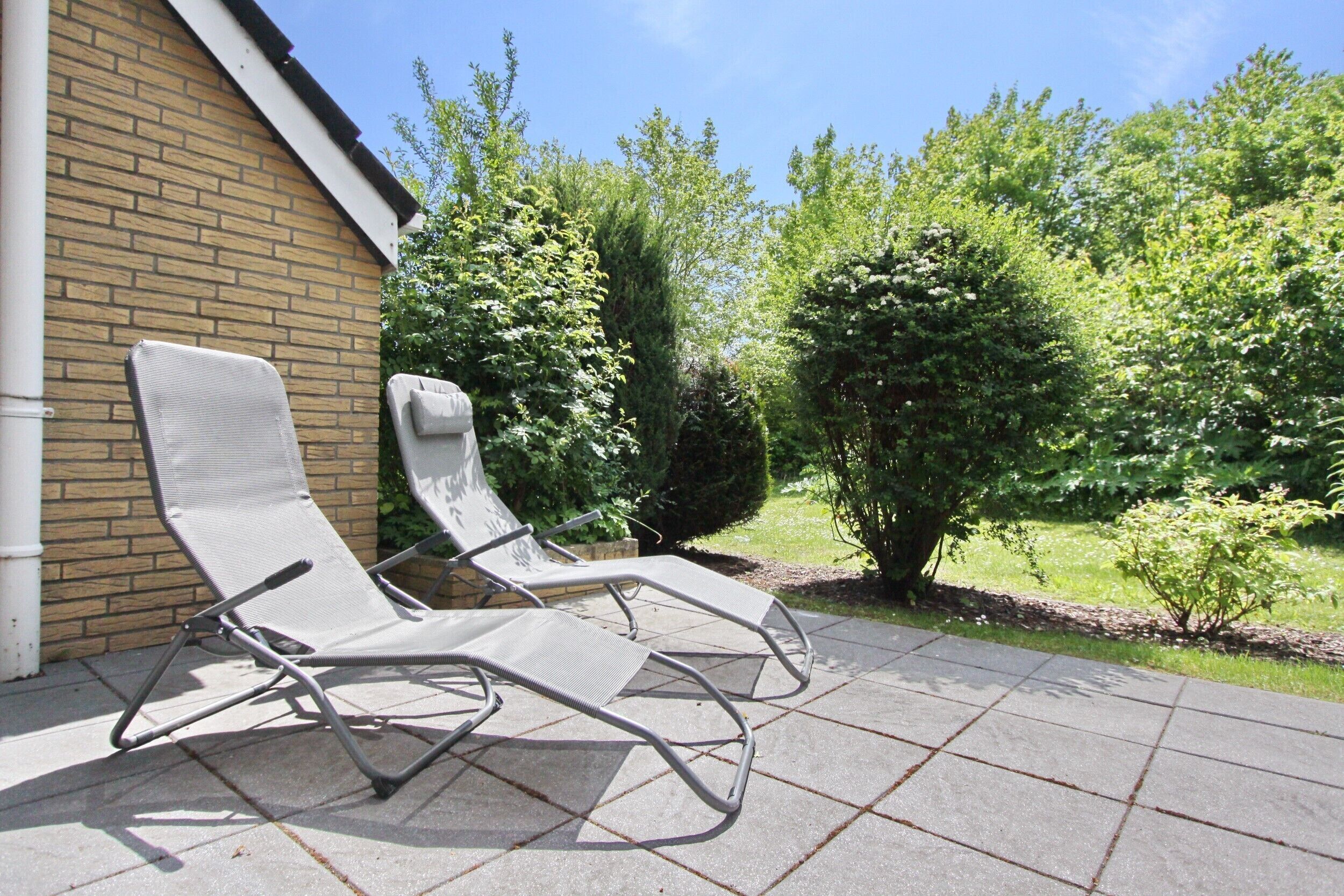 Twee lounge fauteuils op een terras, met een bakstenen muur en groen op de achtergrond.