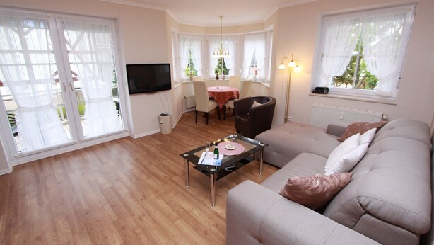 Ein Wohnzimmer mit einer Couch, einem Glastisch, einem Esstisch mit Stühlen und einem an der Wand befestigten Fernseher.