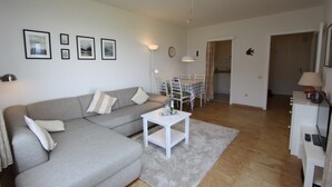 A living room with a grey sofa, a small white table, and a dining area with a table and chairs. - Hanseat II App. 149 Inkl. Wlan (Grömitz)