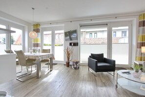 Ein modernes Wohnzimmer mit Essbereich, einem schwarzen Sessel, einem Glastisch und einem an der Wand montierten Fernseher.