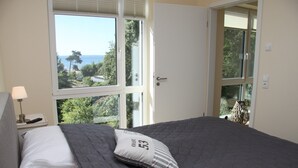 Ein Schlafzimmer mit einem Bett, einem Nachttisch, einer Lampe und Blick ins Freie durch große Fenster.