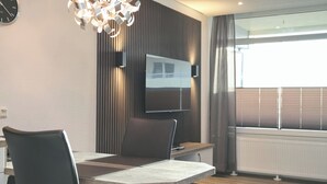 A modern dining area with a table and chairs, a flat-screen TV mounted on a wall, and a large window with blinds. - Grömitz-center II App. 6 Inkl. Wlan (Grömitz)