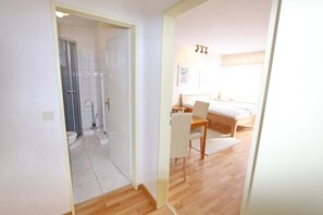 A bedroom with a bed, a desk, and a chair. There is a shower area with a glass door. The room has wooden flooring and white walls.