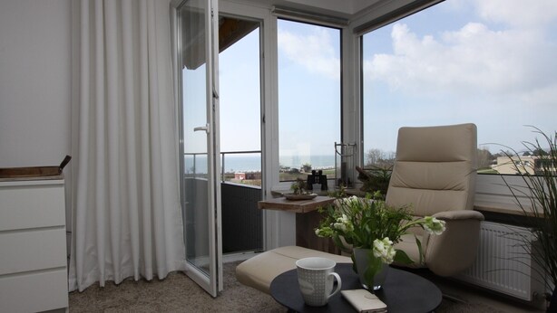 Un balcon avec vue sur la mer, une chaise, une table avec une tasse et un livre, et une plante.