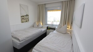 Une petite chambre propre, avec deux lits simples, une fenêtre avec des rideaux et une œuvre d’art accrochée au mur.