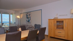 Un comedor con mesa y sillas de madera, un aparador con cristalería, y vistas al mar a través de amplios ventanales.