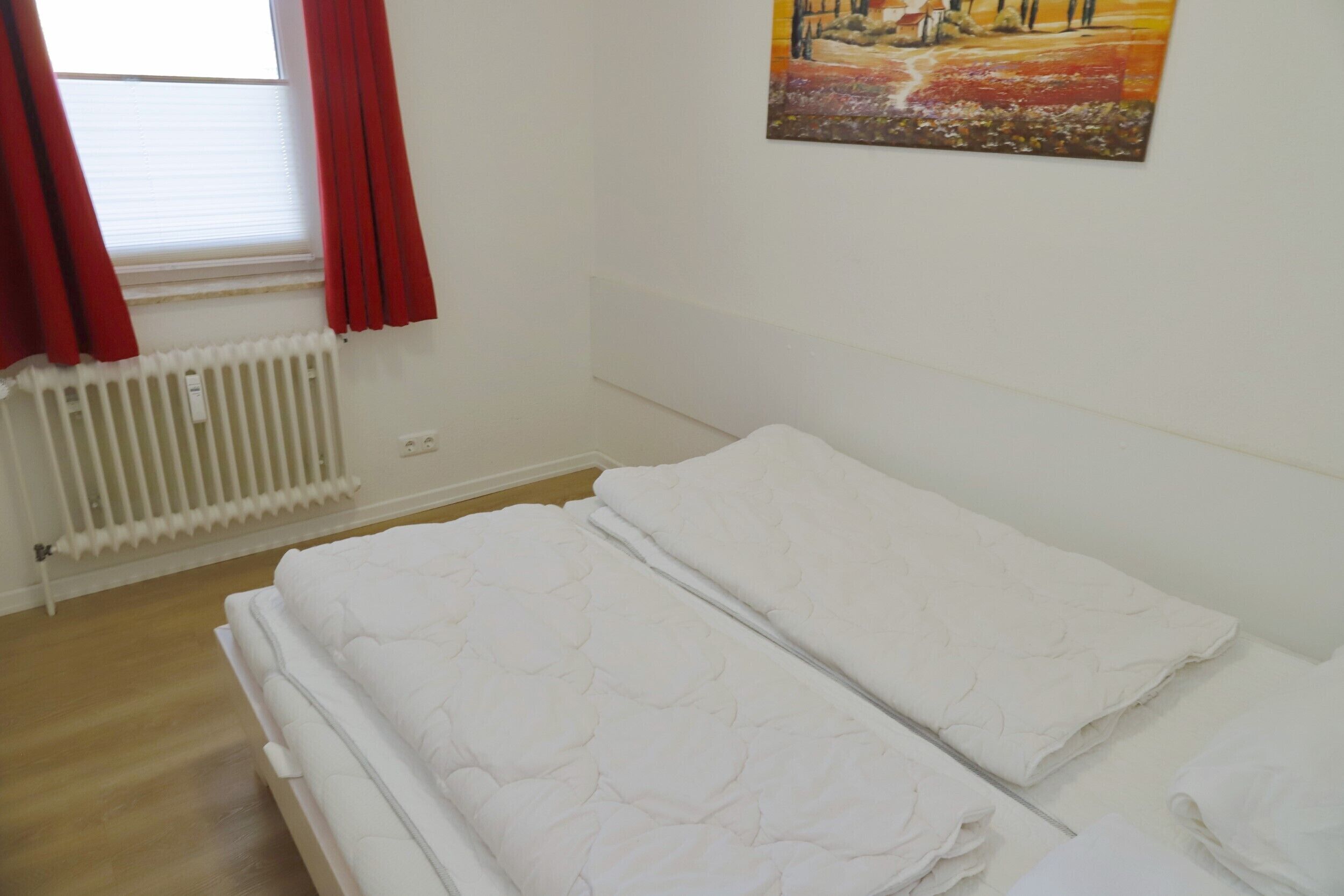 Une chambre avec deux lits, un radiateur, une fenêtre avec des rideaux rouges et un tableau accroché au mur.