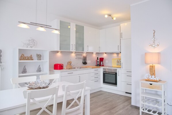 A modern kitchen with white cabinetry, a dining table with chairs, a coffee maker, and a wine rack. - Wiesengrund 12 - 14 Whg. 7 Inkl. Wlan (Grömitz)