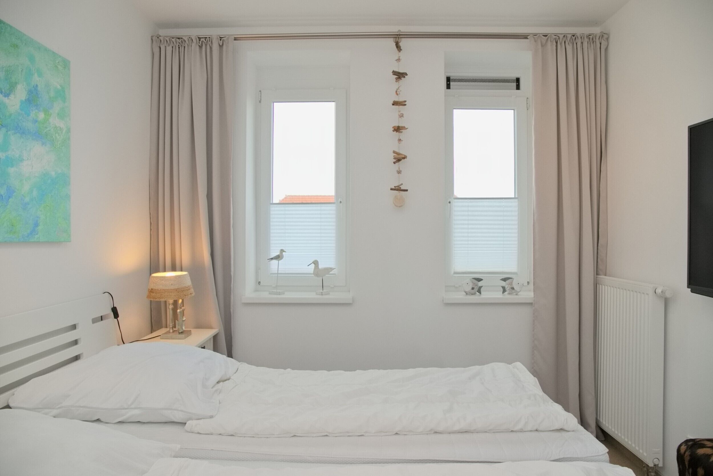 A bedroom with a bed, two windows with curtains, a wall-mounted lamp, and a radiator.