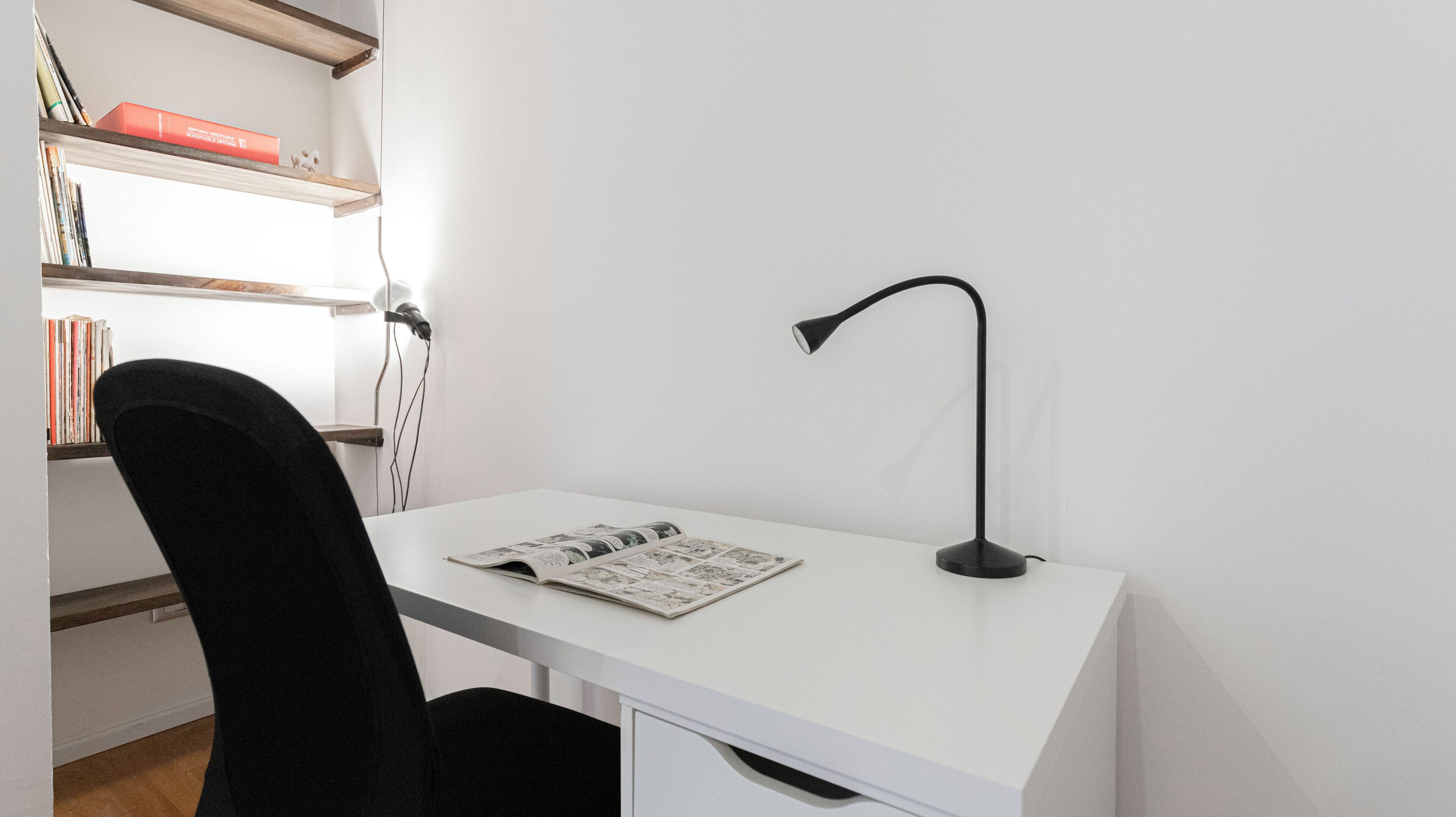 Un bureau blanc avec une lampe de bureau noire, une chaise noire et un magazine posé sur le bureau.