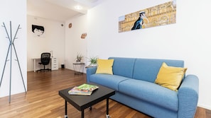 A modern living room with a blue sofa, yellow cushions, a black coffee table with magazines, a tripod stand, a desk with a chair, and a wall with a large painting. - Italianway Easy - Piceno 44 (Milan)