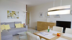 A modern living room with a grey sofa, a glass coffee table, and a wooden dining table with white chairs. There is a TV on a stand and a painting on the wall. - Seeschlößchen App. 113 Inkl. Wlan (Grömitz)