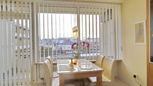 A dining area with a table and chairs, a window with blinds, a hanging light fixture, and a view of buildings outside. - Seeschlößchen App. 113 Inkl. Wlan (Grömitz)