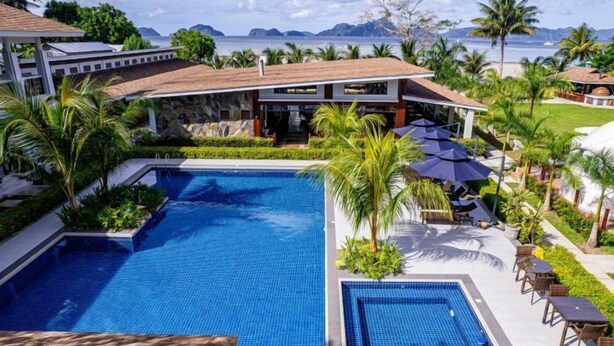 A swimming pool with lounge chairs and umbrellas, surrounded by palm trees and a view of the ocean. - Ora Beach Resort (El Nido)
