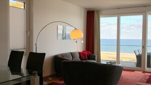 Een moderne woonkamer met een glazen tafel, twee stoelen, een bank, een vloerlamp en uitzicht op het strand.