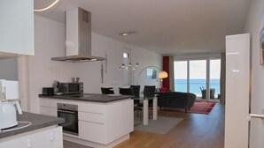 Een moderne keuken met een centraal eiland, een eethoek met een glazen tafel, en uitzicht op de zee door grote ramen.