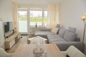 A modern living room with a grey sofa, a wooden coffee table, a TV, and a large window with a view of greenery. - Villa am Meer App. 11 Inkl. Wlan (Grömitz)