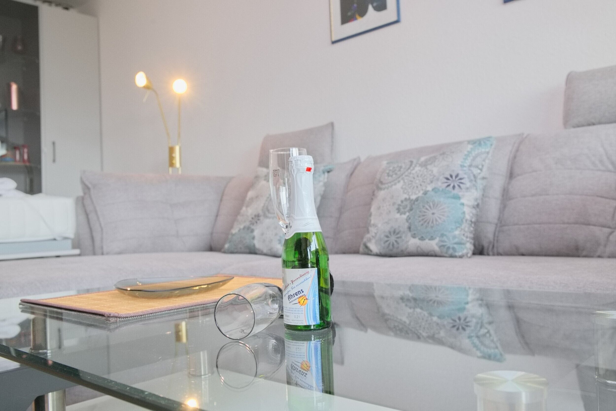 A living room with a glass coffee table, a bottle of green cleaning spray, a pair of glasses, and a cushion on a light-colored sofa.