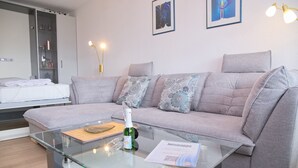 Una sala de estar moderna con una mesa de centro de vidrio, un sofá gris y una cama con ropa de cama blanca.