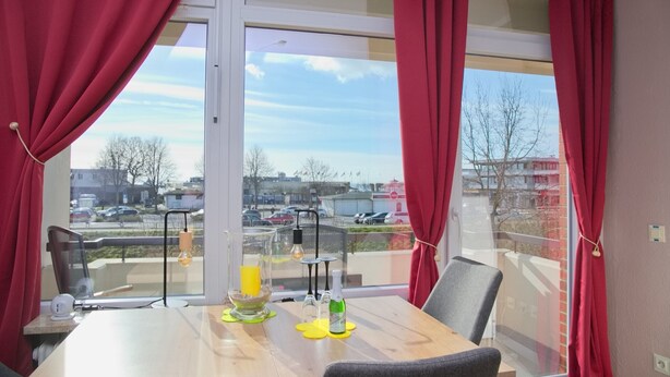 Una habitación con una mesa dispuesta para dos personas, una silla, un ventanal con cortinas rojas y vistas a un estacionamiento y edificios.