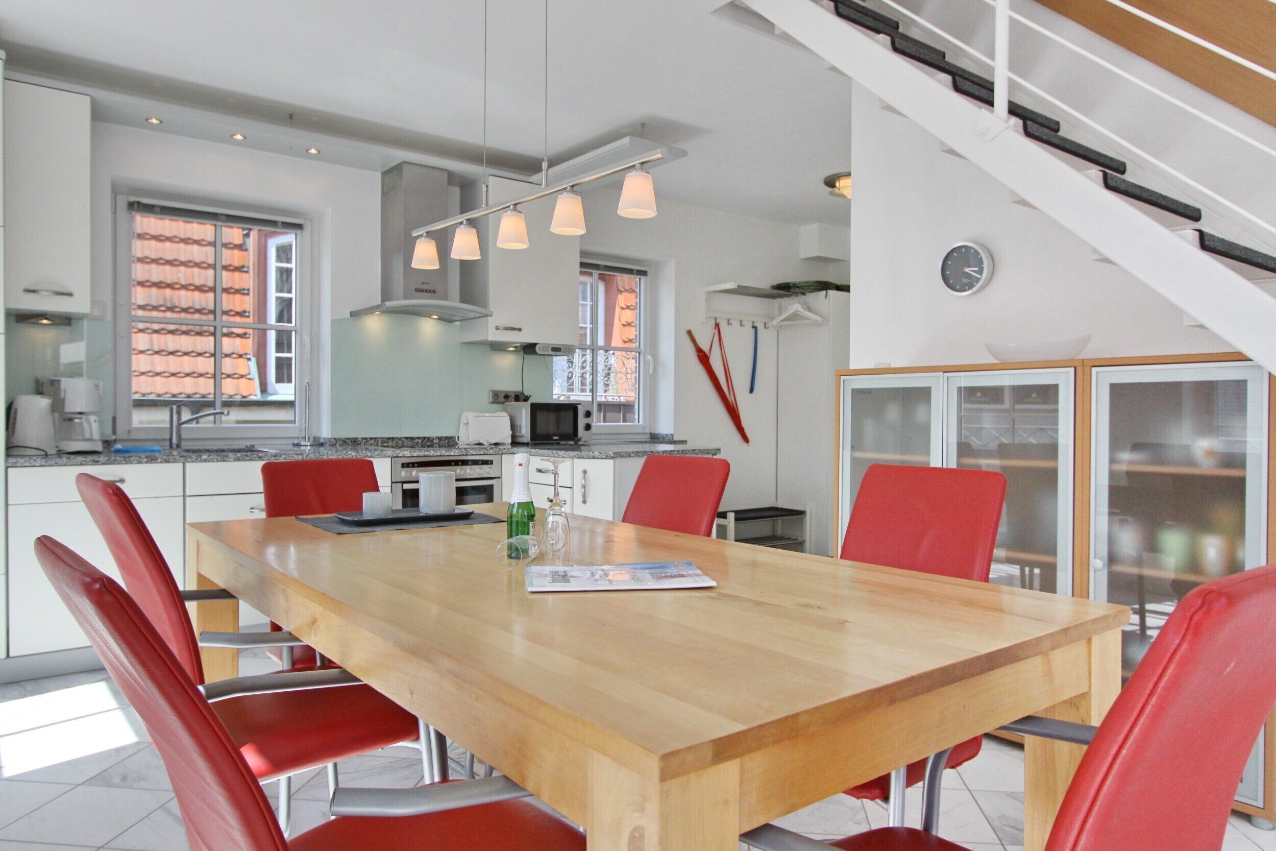 Une cuisine moderne avec une table à manger en bois et des chaises rouges, un escalier et une vue sur l’extérieur.