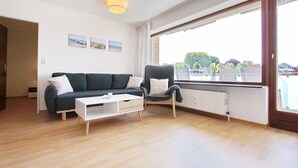 A modern living room with a dark-colored sofa, a white coffee table, and a black armchair. There is a large window providing a view of the outside. - Haus Sonnenblick Whg. 2 Inkl. Wlan (Grömitz)