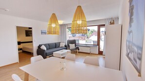 A modern living room with a dining table, sofa, and chairs. There are two hanging light fixtures and a window with a view of buildings. - Haus Sonnenblick Whg. 2 Inkl. Wlan (Grömitz)
