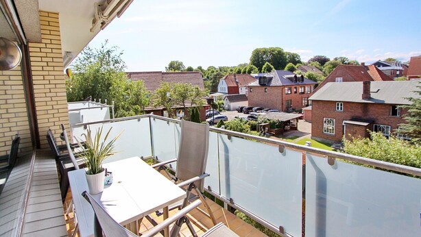 A balcony with a table and chairs, a potted plant, and a view of a residential area with houses and parked cars. - Haus Sonnenblick Whg. 2 Inkl. Wlan (Grömitz)