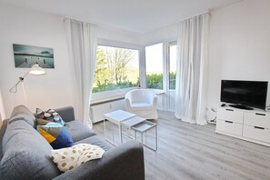 A modern living room with a grey sofa, a small white table, a white chair, a TV on a white cabinet, and large windows with white curtains. - Hanseat II App. 102 Inkl. Strandkorb (Grömitz)