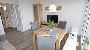A dining area with a wooden table, grey upholstered chairs, a potted plant, and a television on a stand. - Haus Baltic App. 177 Inkl. Wlan (Grömitz)