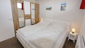 A bedroom with a bed, a nightstand with a lamp, and a wardrobe with a mirror. - Haus Baltic App. 177 Inkl. Wlan (Grömitz)