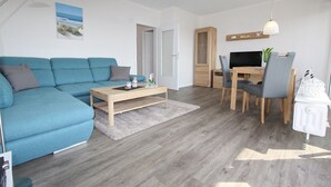 A modern living room with a blue sofa, a wooden coffee table, a dining area with a table and chairs, and a TV on a wooden cabinet. - Haus Baltic App. 177 Inkl. Wlan (Grömitz)