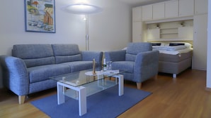 Un soggiorno moderno con un divano grigio, un tavolino in vetro e un letto con testiera bianca.