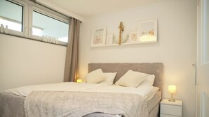 Un dormitorio ordenado con una cama grande, una mesita de noche con lámpara y una ventana con cortinas.