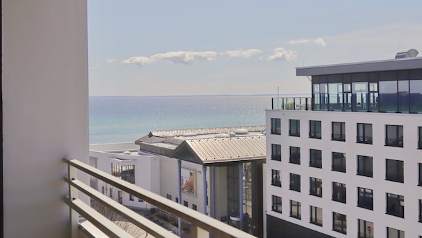 Vista de um varanda sobre uma praia e edifícios modernos, com grandes janelas e céu claro.