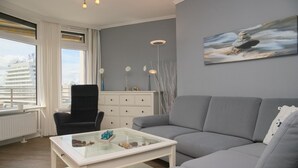 Uma sala de estar moderna com um sofá cinzento, uma mesa de centro de vidro e uma cadeira preta. Há uma pintura na parede e uma janela com vista para edifícios.